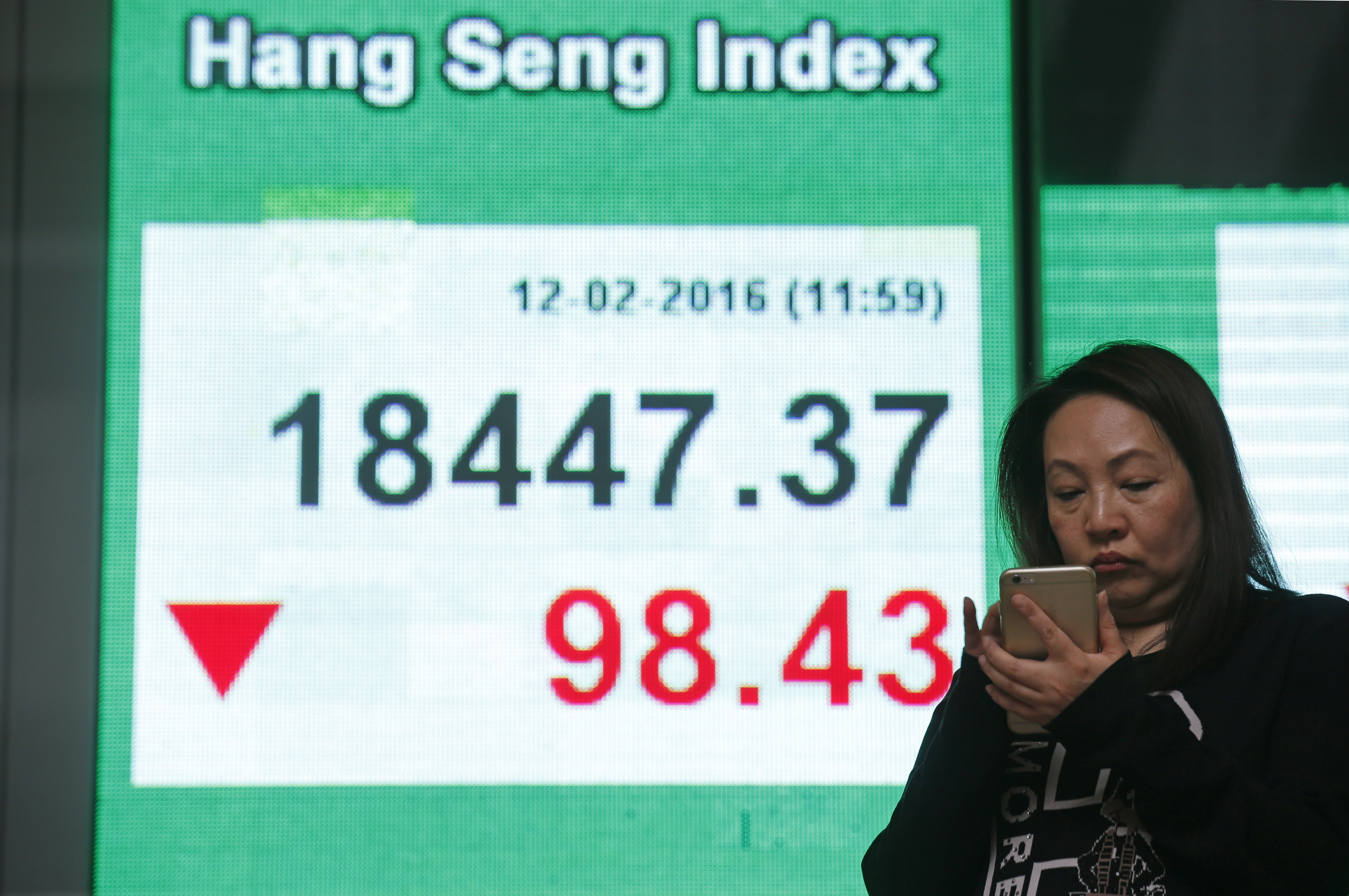 A woman uses her smartphone in front of an electronic board showing the Hong Kong index at a bank in Hong Kong, Friday, Feb. 12, 2016. Japan’s main stock index dived Friday, leading other Asian markets lower, after a sell-off in banking shares roiled investors in the U.S. and Europe. (AP Photo/Kin Cheung)