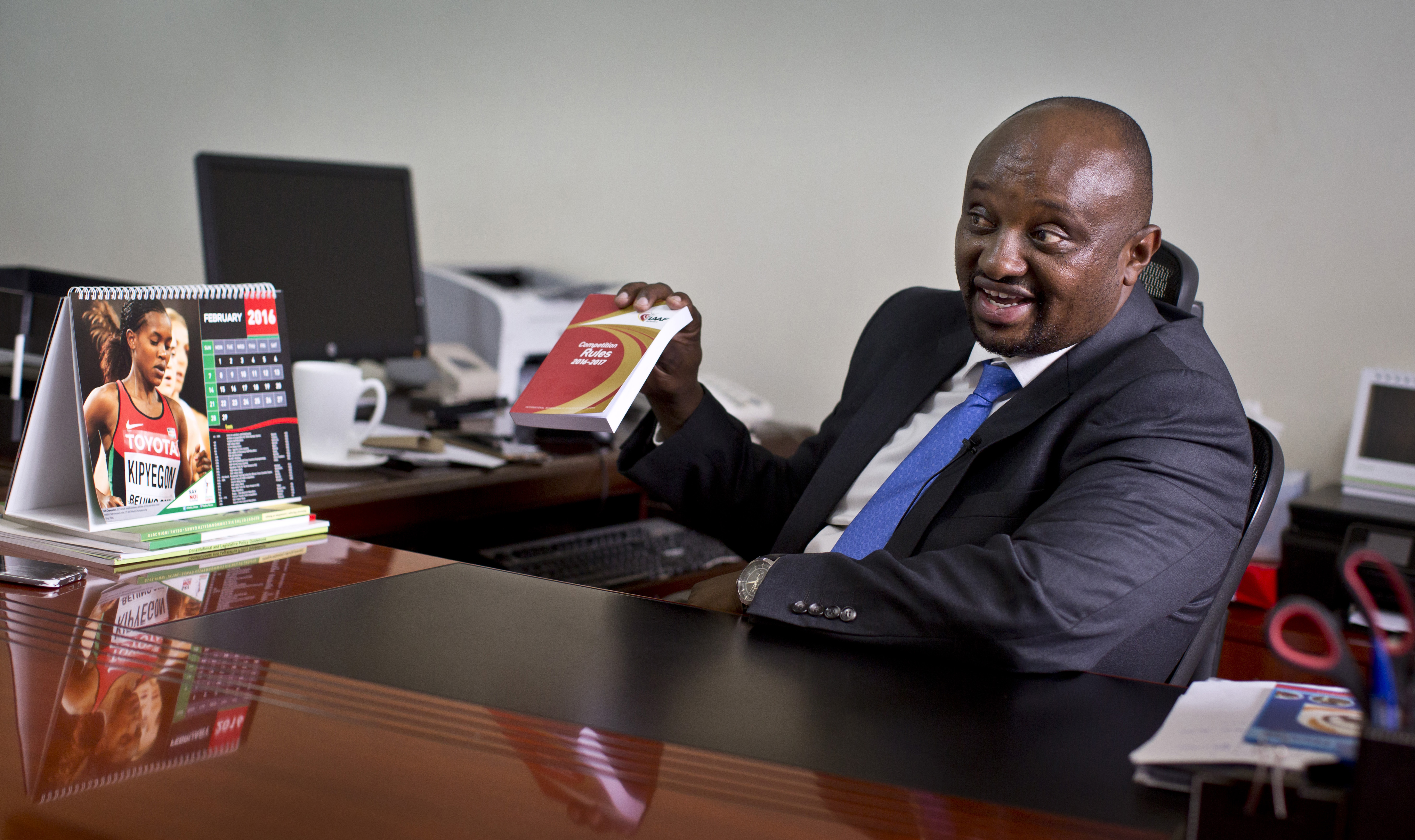 FILE---In this file photo taken Monday, Feb. 8, 2016, Athletics Kenya CEO Isaac Mwangi holds up a copy of the IAAF Competition Rules book, to illustrate his denial that Athletics Kenya would have the power to shave time off athletes’ bans, during an interview with The Associated Press at his office in Nairobi, Kenya. The chief executive of the track and field governing body in running powerhouse Kenya has temporarily stepped aside after two athletes alleged in an interview with The Associated Press that he asked them for a bribe to reduce their bans for doping. (AP Photo/Ben Curtis, File)