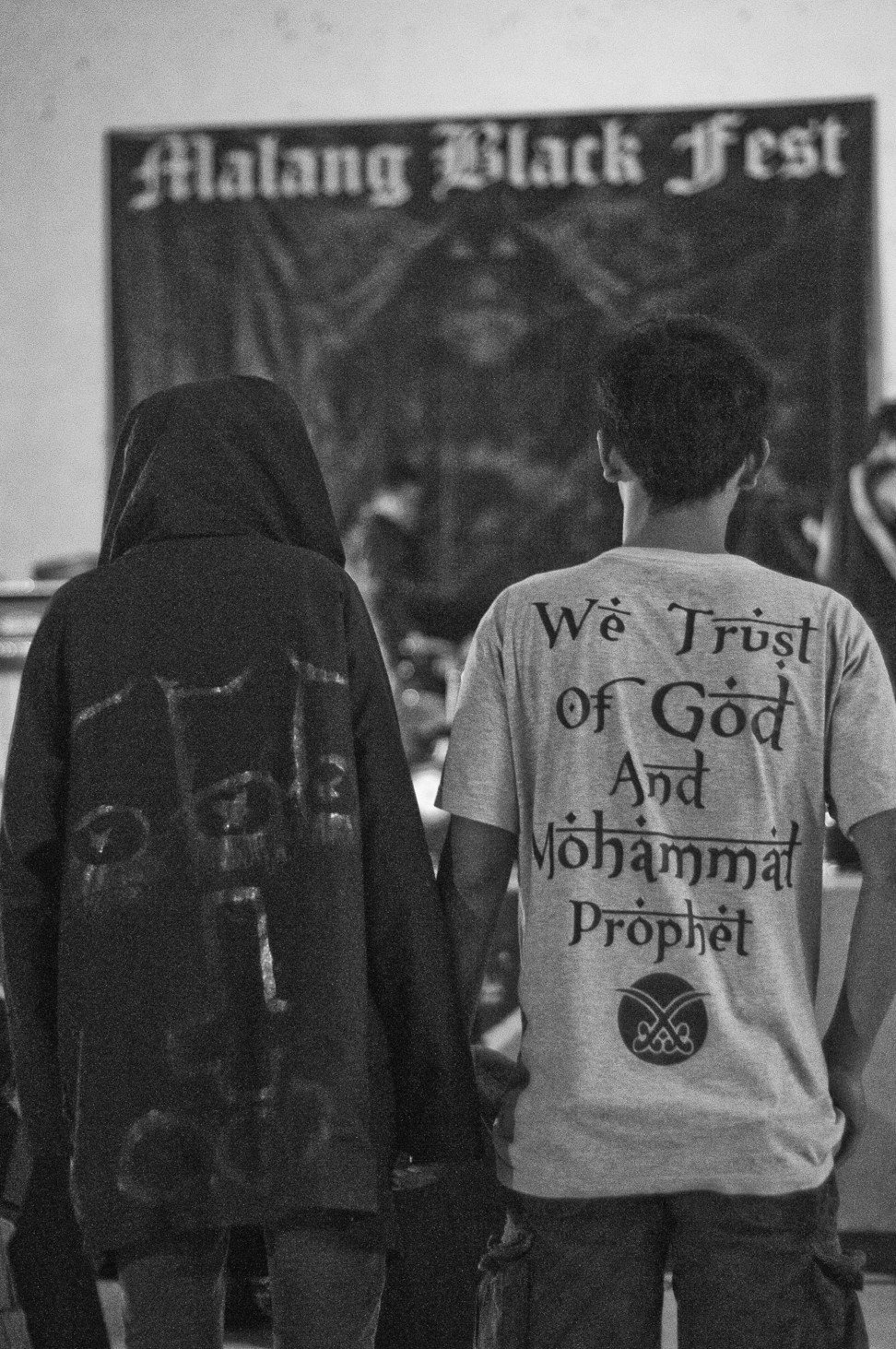 At a black metal show in Malang, East Java, a young Indonesian (left) wearing a cowl with 666 and an inverted crucifix stands next to another man wearing a T-shirt that praises god and the prophet Muhammad. Photo: Gede Adhiputra